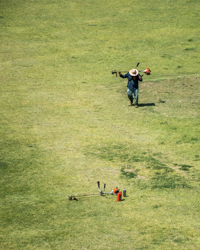 High Angle Shot of a Man Walking on a Grass Field with a Grass Trimmer